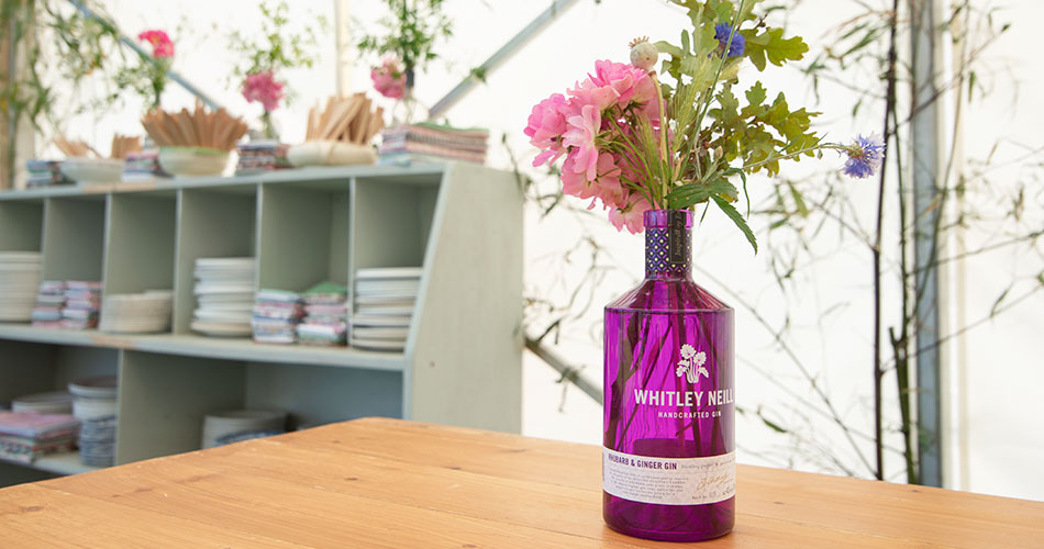 A pink glass bottle of gin being used for a flower vase with a lovely bouquet of summer flowers and a dresser with vintage plates in the background